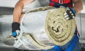 Budowlaniec w rękawicach niesie zrolowaną wełnę mineralną, owiniętą folią aluminiową.