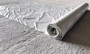 Biała folia budowlana rozwinięta na szarej, pylistej podłodze. Rolka folii leży po prawej stronie, tworząc cień.
