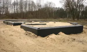 Fundament domu w piasku, z czarnym lepikiem izolującym ściany fundamentowe. W tle las.