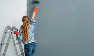 Dziewczyna w kratkę i pomarańczowych rękawiczkach maluje ścianę na szaro. Obok stoi drabina. Zastanawiasz się, ile farby na m2 zużyjesz?