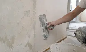 Ręka nakłada gładź polimerową na ścianę za pomocą kielni. Widać starą, łuszczącą się warstwę tynku i wiadro z masą.