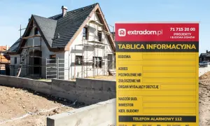 Tablica informacyjna budowy domu w trakcie prac, z widocznym rusztowaniem i fundamentami. Na tablicy dane projektu i numery alarmowe.