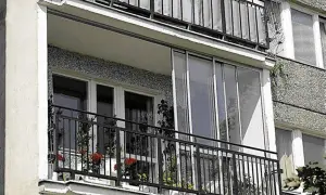Zabudowa balkonu w bloku z przeszklonymi drzwiami i metalową balustradą. Na balkonie stoją doniczki z kwiatami.