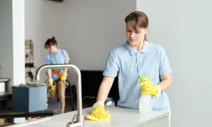 Dwie kobiety w niebieskich koszulkach sprzątają mieszkanie. Jedna wyciera blat, druga odkurza. Zastanawiasz się, ile kosztuje sprzątanie mieszkania?