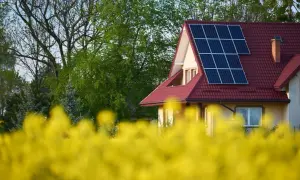 Dom z instalacją PV na czerwonym dachu, otoczony kwitnącym rzepakiem i zielenią drzew.
