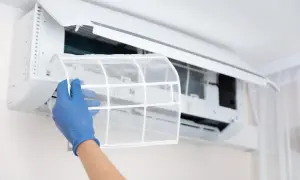 Ręka w niebieskiej rękawiczce wyjmuje filtr z klimatyzatora domowego. Dbanie o czystość to podstawa.