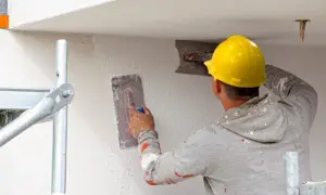 Pracownik w żółtym kasku wykonuje tynkowanie elewacji, używając kielni do nakładania masy.