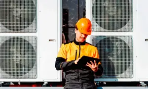 Technik w pomarańczowym kasku sprawdza dane na tablecie przy jednostkach zewnętrznych pompy ciepła. Dofinansowanie do pompy ciepła może być w zasięgu ręki!