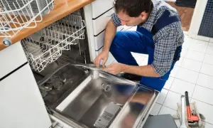 Fachowiec w niebieskim kombinezonie dokonuje podłączenia zmywarki, pracując przy jej wnętrzu. Obok leżą narzędzia.