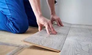 Ręce układają szare panele podłogowe, pokazując, jak kłaść panele. Widać niebieskie spodnie i fragment ściany.