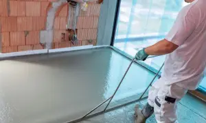 Pracownik wylewa i wyrównuje świeżą posadzkę za pomocą narzędzi. Ściana z cegły i okno w tle.