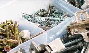 Pudełko z różnymi śrubkami, wkrętami i elementami metalowymi. Widać złote i srebrne śrubki, gwoździe, a także metalowe zawiasy.