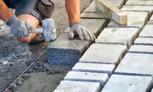 Brukarz układa kamienne płyty, używając młotka. Widać mokry, świeży beton, a obok suche płyty, sugerujące odpowiednie suchy beton proporcje.
