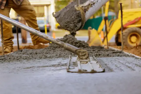 Pracownik budowlany wyrównuje świeży beton, który właśnie wylano z betoniarki. To idealny moment, by sprawdzić, jaki beton na chudziaka będzie najlepszy. W tle widać koparkę.