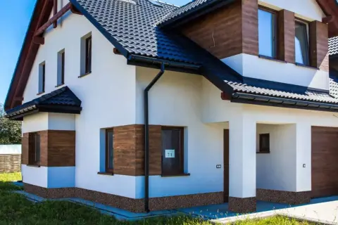 Nowoczesny dom z białym tynkiem, drewnianymi akcentami i ciemnym dachem. Uwagę przyciąga cokół z płytek imitujących kamień.