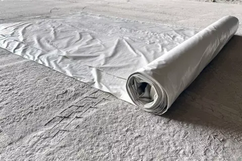 Biała folia budowlana rozwinięta na szarej, pylistej podłodze. Rolka folii leży po prawej stronie, tworząc cień.