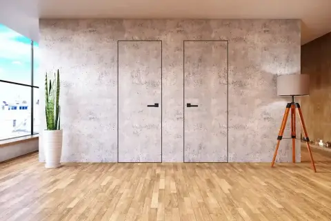 Dwa minimalistyczne, **drzwi ukryte w ścianie** z betonu architektonicznego, z czarnymi uchwytami. Obok stoi lampa i roślina.