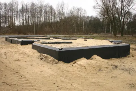 Fundament domu w piasku, z czarnym lepikiem izolującym ściany fundamentowe. W tle las.