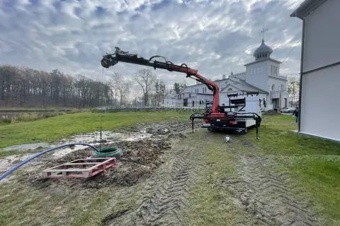 Prace przy studni głębinowej: dźwig z ramieniem pracuje nad wykończeniem, w tle budynek.