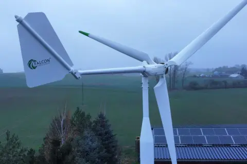 Biała elektrownia wiatrowa Falcon z zielonymi akcentami na tle zielonych pól i nieba. Obok widoczne panele słoneczne na dachu.