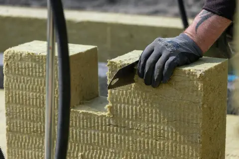 Ręka w rękawicy tnie piłą wełnę skalną 10. Materiał izolacyjny jest cięty na budowie.
