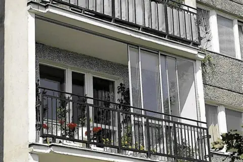 Zabudowa balkonu w bloku z przeszklonymi drzwiami i metalową balustradą. Na balkonie stoją doniczki z kwiatami.