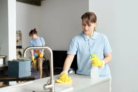 Dwie kobiety w niebieskich koszulkach sprzątają mieszkanie. Jedna wyciera blat, druga odkurza. Zastanawiasz się, ile kosztuje sprzątanie mieszkania?