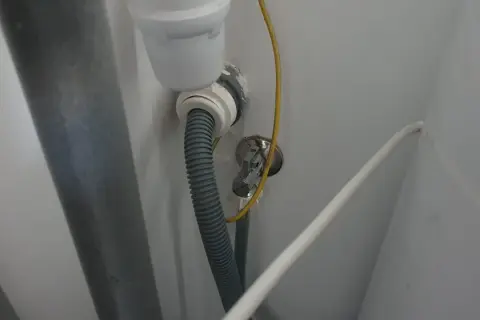 Szara rura odpływowa pralki podłączona do syfonu pod zlewem. Widoczny żółty przewód i biały kabel.