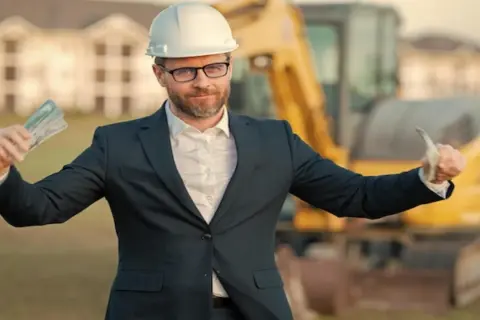 Kierownik budowy w kasku i garniturze pokazuje pieniądze. Zastanawiasz się, ile kosztuje kierownik budowy?