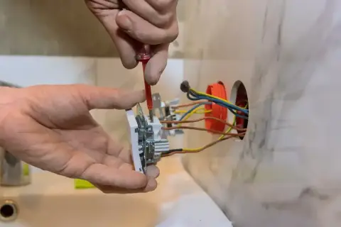 Instalator przykręca gniazdko, pokazując, jak podłączyć gniazdko. Widać przewody elektryczne i narzędzia.