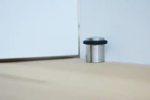 Metalowy odbojnik z czarną gumką przy ścianie, chroniący drzwi przed uderzeniem.