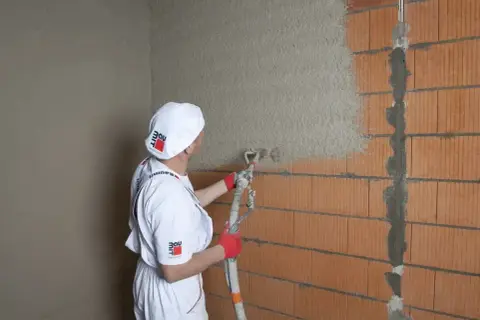 Tynkarz w białym ubraniu i czapce z logo Baumit nakłada tynk agregatem na ścianę z cegły.
