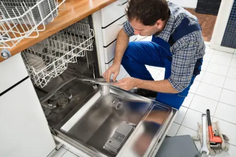 Fachowiec w niebieskim kombinezonie dokonuje podłączenia zmywarki, pracując przy jej wnętrzu. Obok leżą narzędzia.