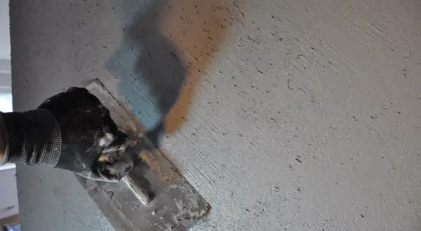 Ręka w rękawicy wygładza świeżo nałożony tynk, pokazując, jak zrobić beton. Cień narzędzia rzuca się na szarą, chropowatą powierzchnię.