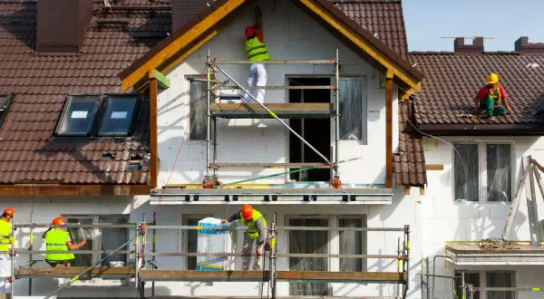 Pracownicy na rusztowaniach prowadzą termomodernizację domu, docieplając ściany i dach.