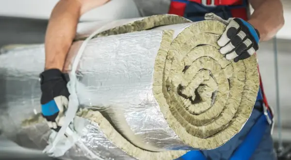 Budowlaniec w rękawicach niesie zrolowaną wełnę mineralną, owiniętą folią aluminiową.