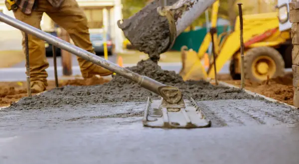 Pracownik budowlany wyrównuje świeży beton, który właśnie wylano z betoniarki. To idealny moment, by sprawdzić, jaki beton na chudziaka będzie najlepszy. W tle widać koparkę.