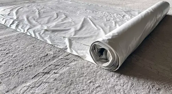 Biała folia budowlana rozwinięta na szarej, pylistej podłodze. Rolka folii leży po prawej stronie, tworząc cień.