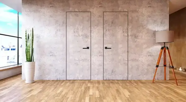 Dwa minimalistyczne, **drzwi ukryte w ścianie** z betonu architektonicznego, z czarnymi uchwytami. Obok stoi lampa i roślina.