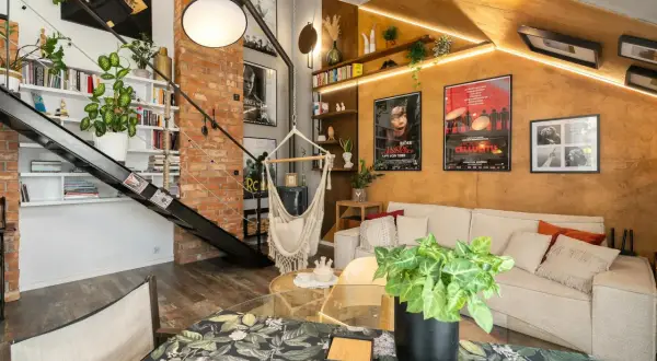 Salon w stylu loftowym z ceglaną ścianą, metalowymi schodami, wiszącym fotelem i przytulną kanapą.
