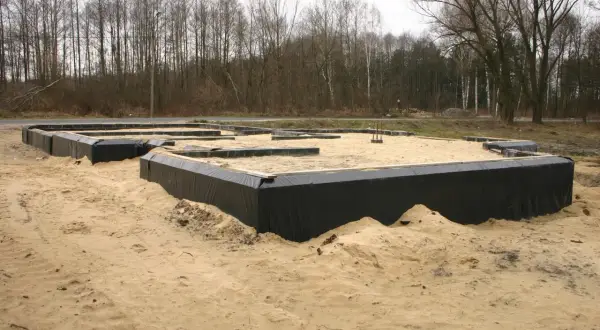 Fundament domu w piasku, z czarnym lepikiem izolującym ściany fundamentowe. W tle las.