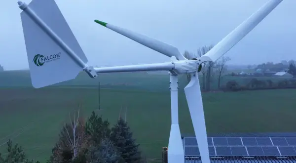 Biała elektrownia wiatrowa Falcon z zielonymi akcentami na tle zielonych pól i nieba. Obok widoczne panele słoneczne na dachu.