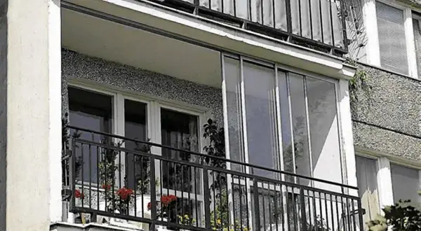 Zabudowa balkonu w bloku z przeszklonymi drzwiami i metalową balustradą. Na balkonie stoją doniczki z kwiatami.
