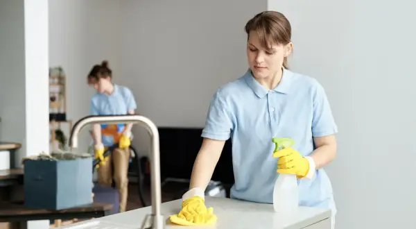 Dwie kobiety w niebieskich koszulkach sprzątają mieszkanie. Jedna wyciera blat, druga odkurza. Zastanawiasz się, ile kosztuje sprzątanie mieszkania?