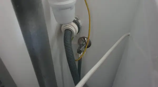 Szara rura odpływowa pralki podłączona do syfonu pod zlewem. Widoczny żółty przewód i biały kabel.