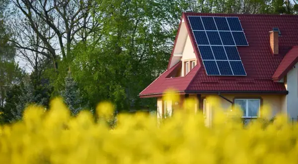 Dom z instalacją PV na czerwonym dachu, otoczony kwitnącym rzepakiem i zielenią drzew.