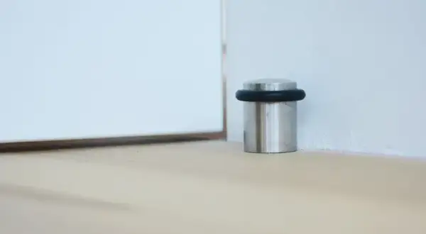 Metalowy odbojnik z czarną gumką przy ścianie, chroniący drzwi przed uderzeniem.