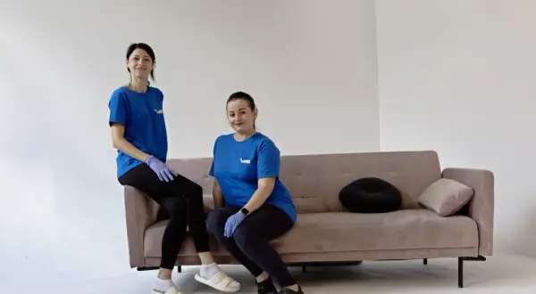Dwie panie w niebieskich koszulkach i rękawiczkach siedzą na kanapie. Zastanawiasz się, ile kosztuje sprzątanie domu raz w tygodniu?