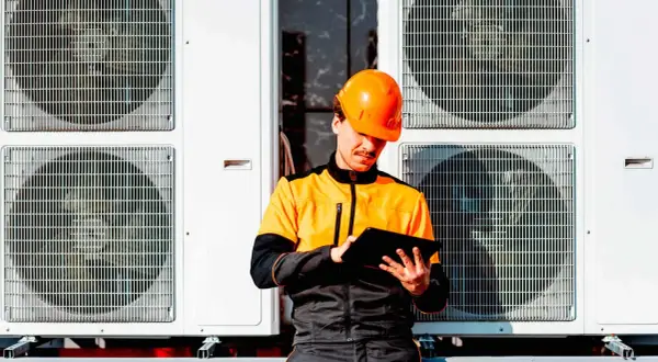 Technik w pomarańczowym kasku sprawdza dane na tablecie przy jednostkach zewnętrznych pompy ciepła. Dofinansowanie do pompy ciepła może być w zasięgu ręki!