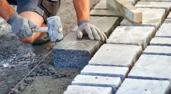 Brukarz układa kamienne płyty, używając młotka. Widać mokry, świeży beton, a obok suche płyty, sugerujące odpowiednie suchy beton proporcje.